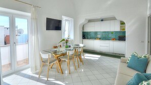 A bright, open-plan living and dining area with a large window, a flat-screen TV, and a kitchen with white cabinets and a green backsplash.