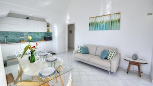 A modern kitchen with white cabinets, a dining area with a glass table, and a living room with a sofa and a painting on the wall.