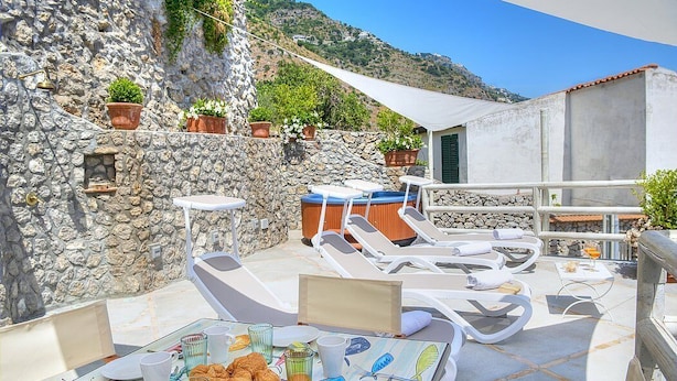 A rooftop terrace with stone walls, lounge chairs, a hot tub, and a table set for breakfast.