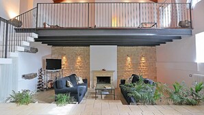 Un grand salon doté d’une cheminée, de deux canapés, d’une table basse et d’un téléviseur. Il y a un balcon avec une balustrade et un mur en pierre.