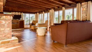 A spacious living area with a brick fireplace, wooden ceiling beams, and large windows offering a view of palm trees and mountains.