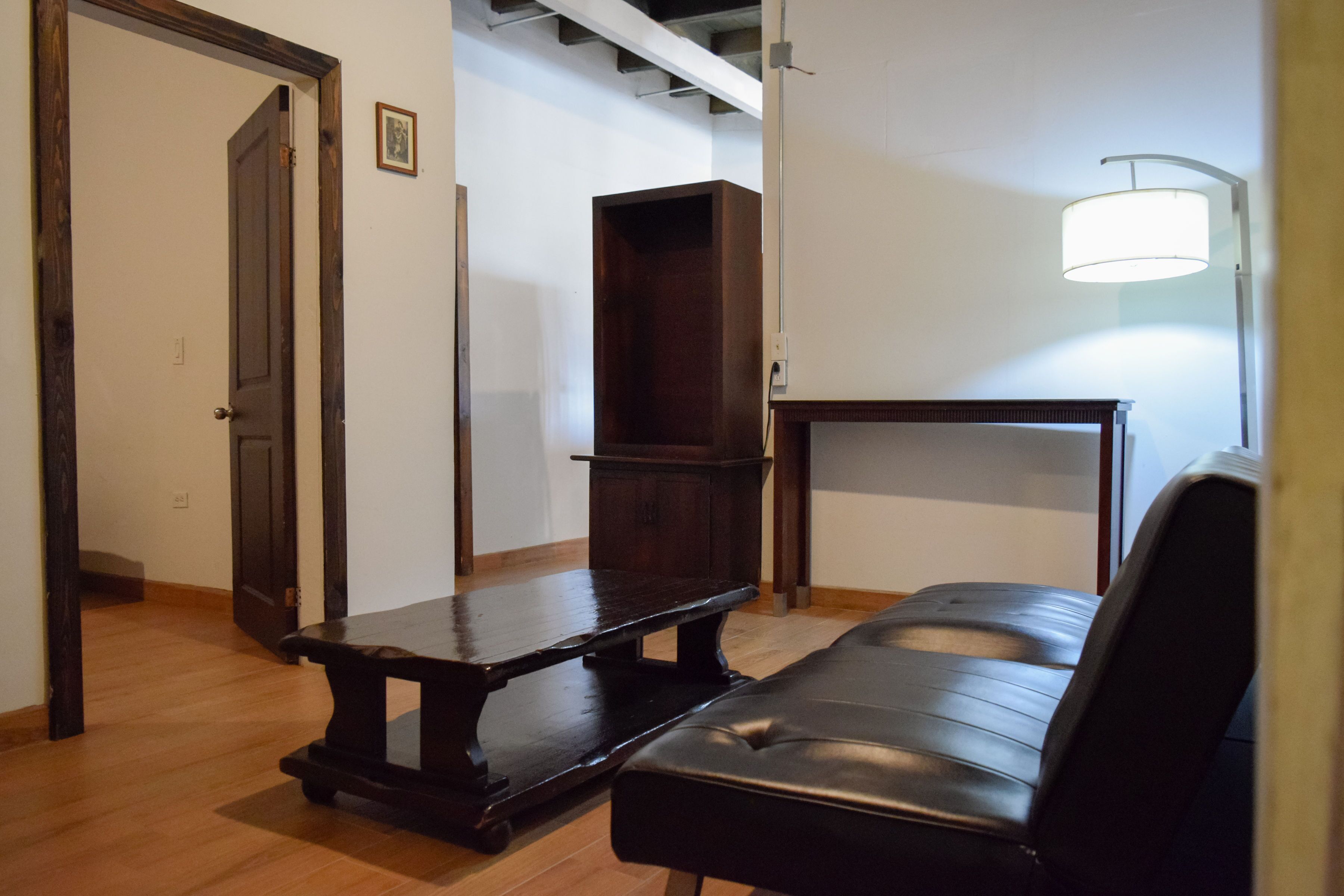 A room with a wooden bench, a black leather sofa, a wooden cabinet, and a wall-mounted lamp.
