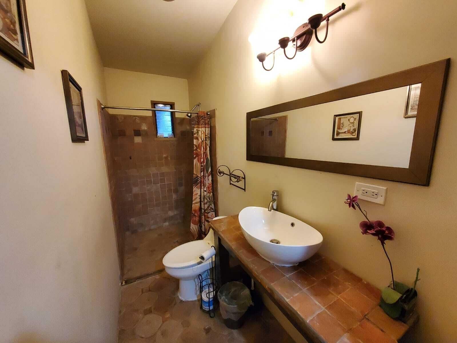 Casa de banho com chuveiro, duas pias, sanita e um espelho, além de uma pequena janela.