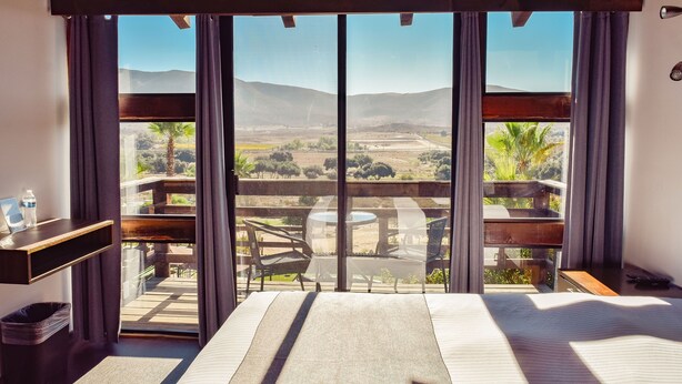 A room with a bed, a small table, a chair, and a view of a balcony with a table and chairs.