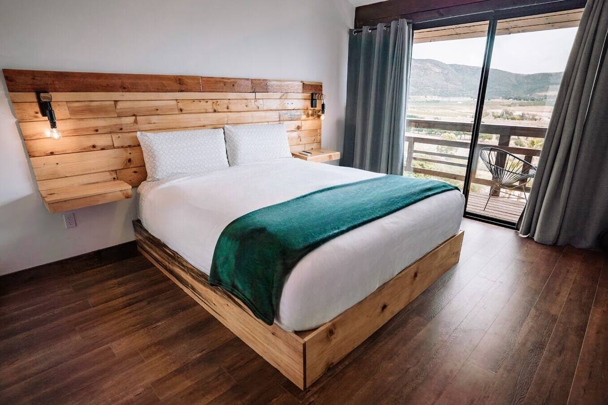 Une chambre moderne avec une tête de lit en bois, un grand lit avec une literie blanche et un plaid vert, et une vue sur un paysage montagneux par une porte-fenêtre coulissante.