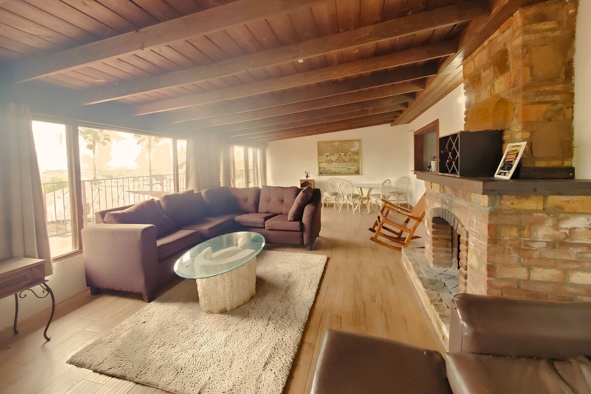 A cozy living room with a stone fireplace, wooden ceiling beams, a glass coffee table, and a seating area with a sofa and chairs.