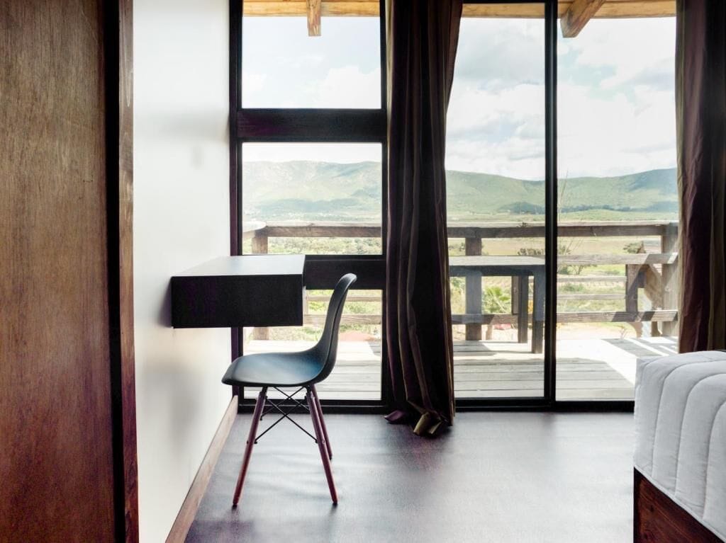 A bedroom with a bed, a desk, and a chair. There is a large window with a view of hills.