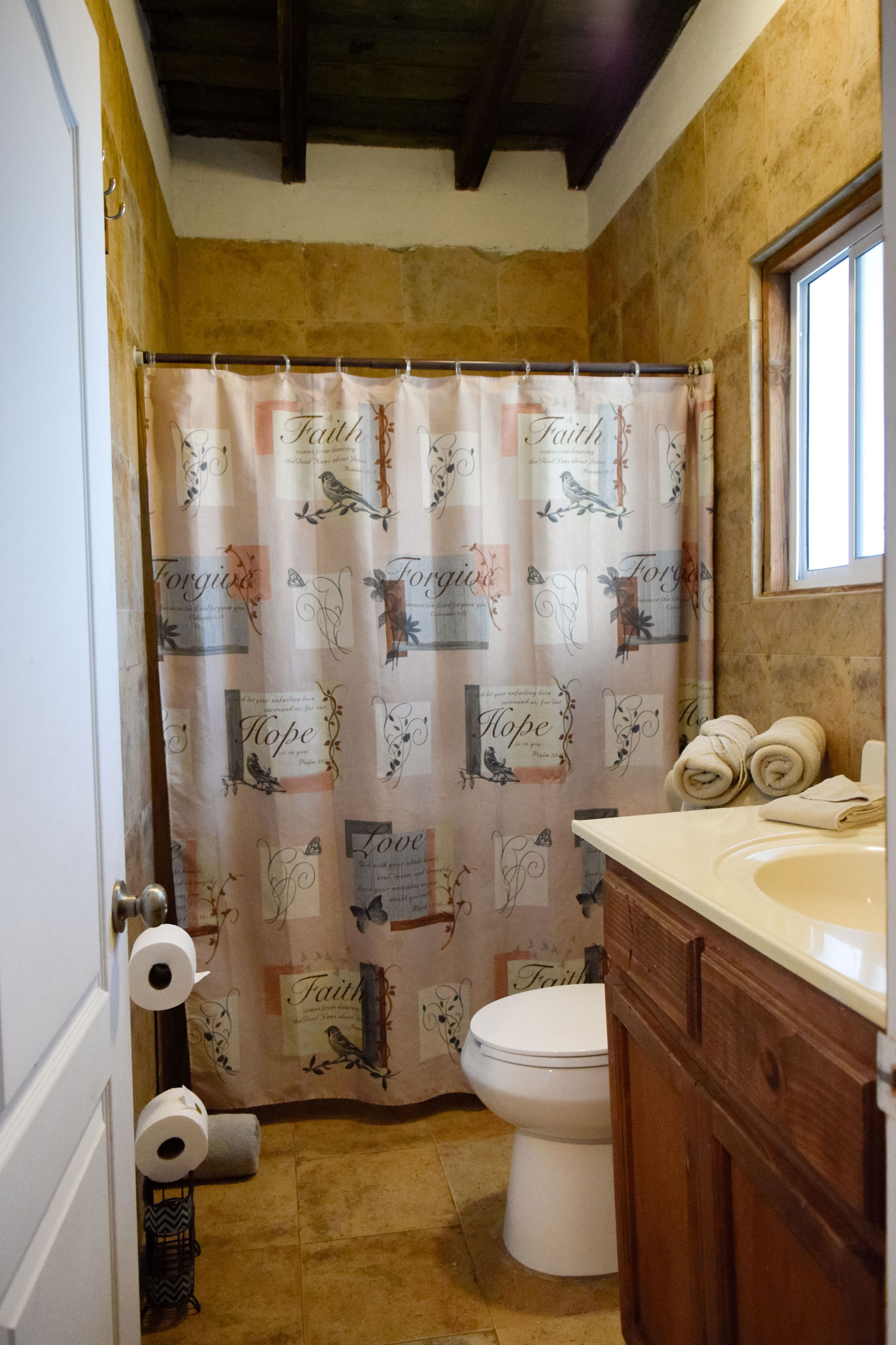 A bathroom with a shower curtain featuring religious-themed prints, a toilet, a sink with towels, and a window.