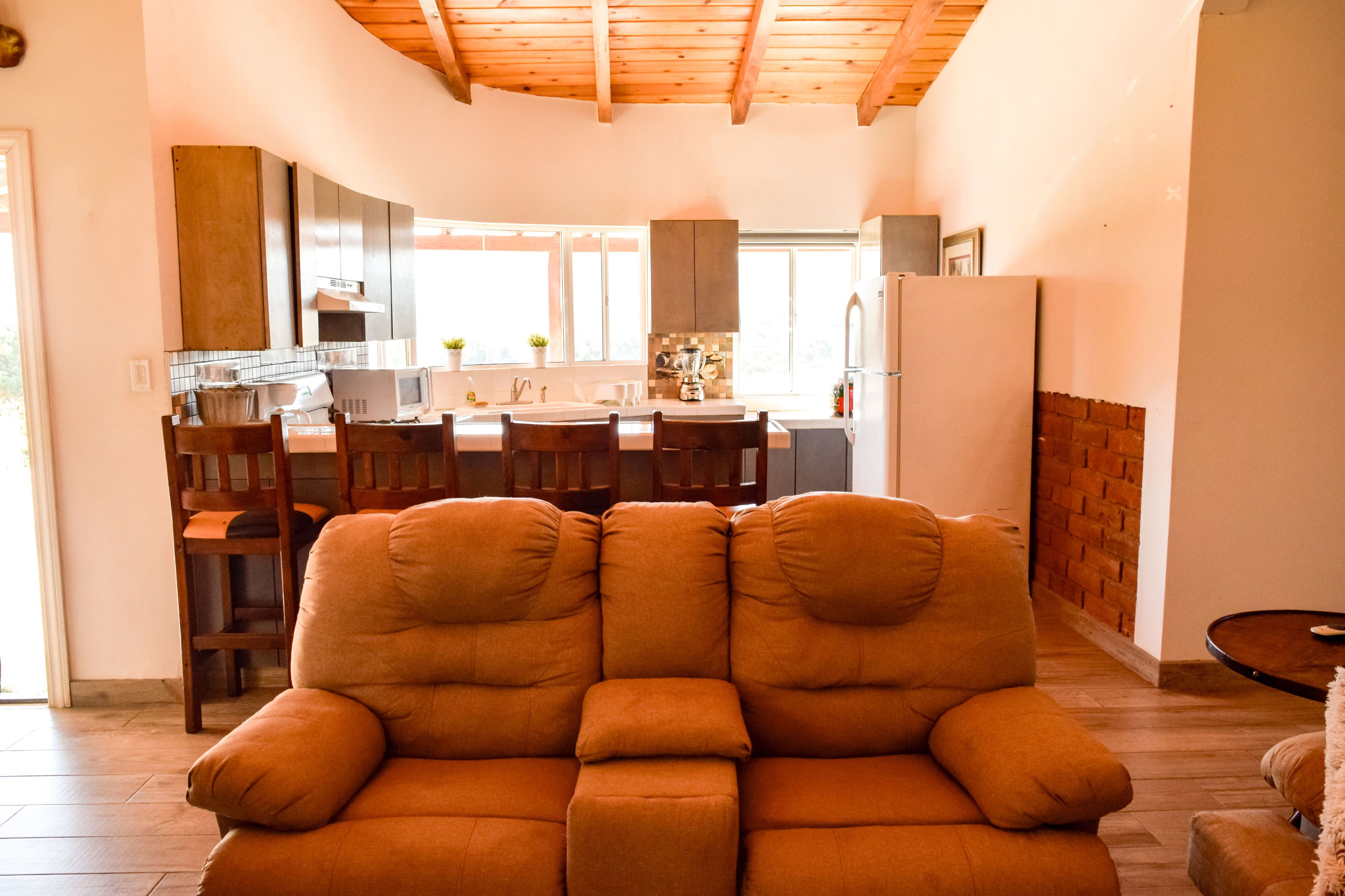 Una sala de estar moderna con sofá, comedor con sillas, cocina equipada y un gran ventanal que proporciona luz natural.