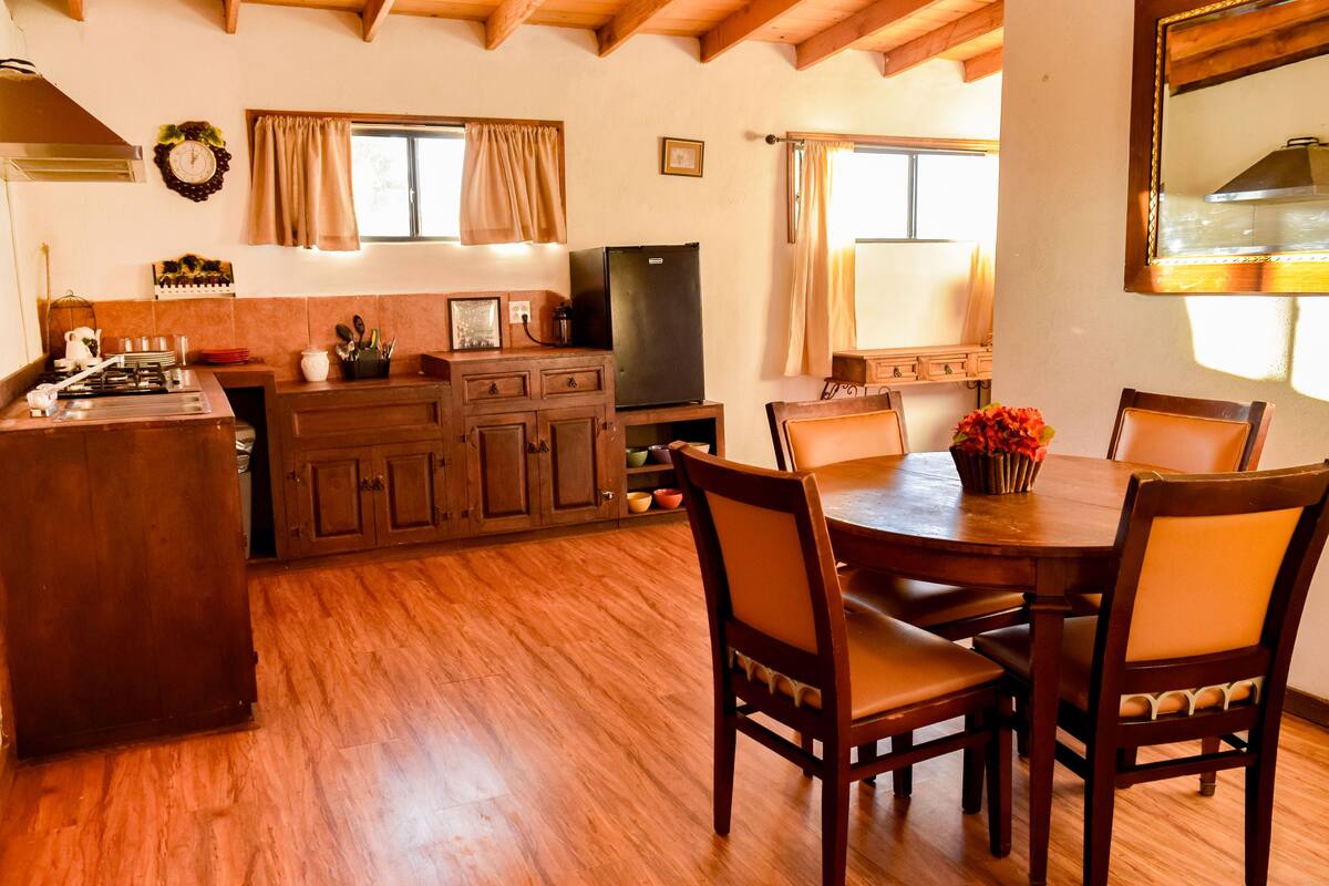 Une table de salle à manger en bois, avec six chaises, une cuisine dotée de meubles en bois, d’un réfrigérateur et d’une cuisinière.
