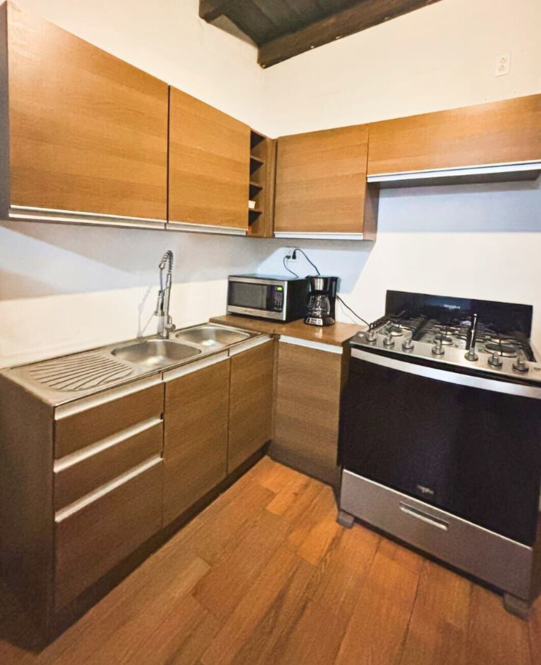 Una cocina compacta con armarios de madera, un lavamanos de acero inoxidable, un microondas, una cafetera y una estufa a gas.