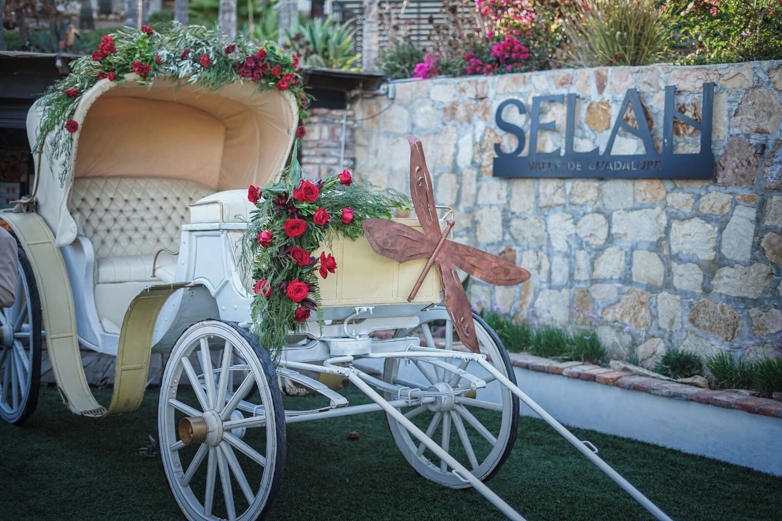 A vintage carriage with floral decorations parked in front of a stone wall with a sign reading "SELAH VILLA DE GUADALUPE".