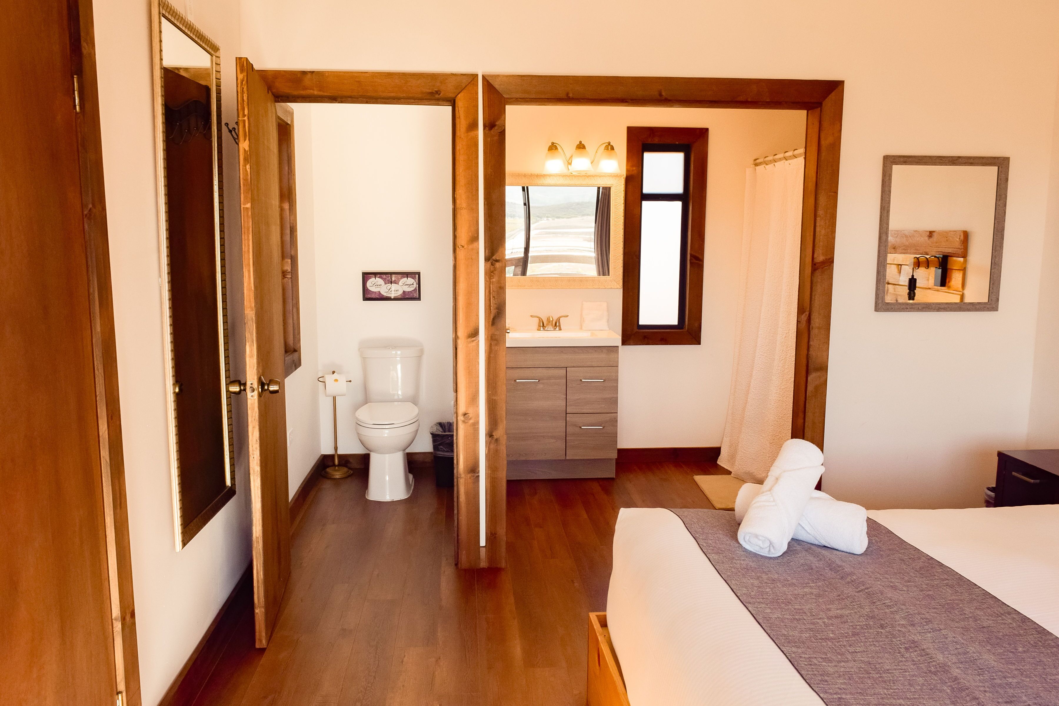 Habitación de hotel con piso de madera, cama con manta gris, baño con inodoro, lavamanos y espejo.