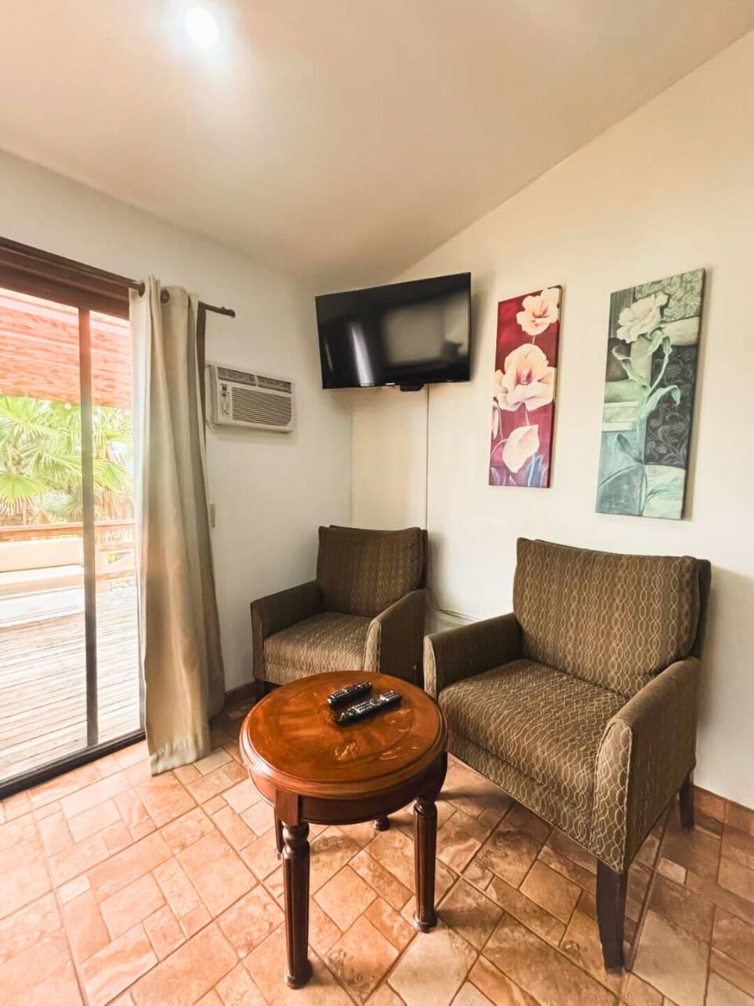 A room with two armchairs, a round wooden table with two remotes, a flat-screen TV mounted on the wall, and a sliding glass door leading to an outdoor area.