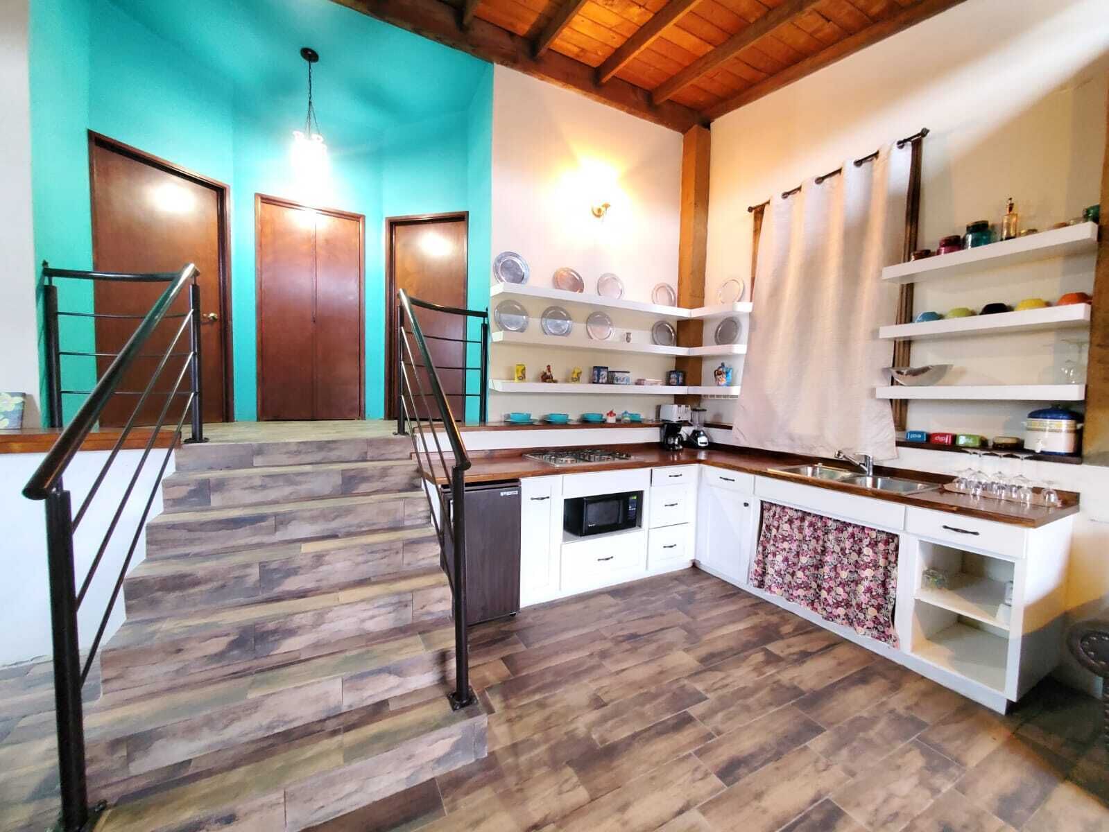 Cozinha com piso de madeira, armários brancos, forno embutido e micro-ondas. Prateleiras com louças e luminária pendente.