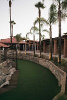 A courtyard with a green artificial turf, palm trees, stone walls, and traditional architecture with red-tiled roofs.