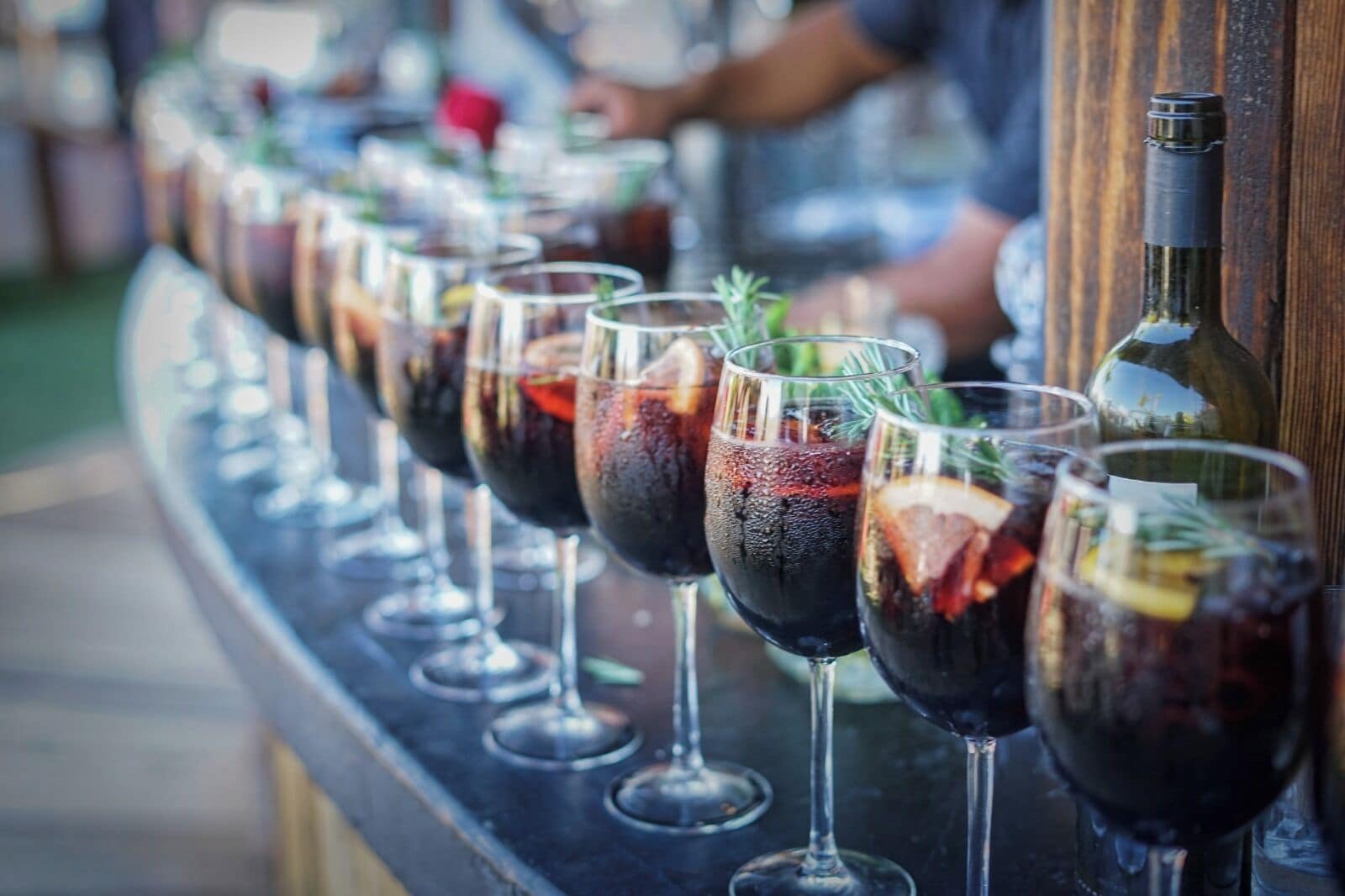 A row of wine glasses filled with red wine, garnished with herbs and citrus slices, displayed on a bar counter.