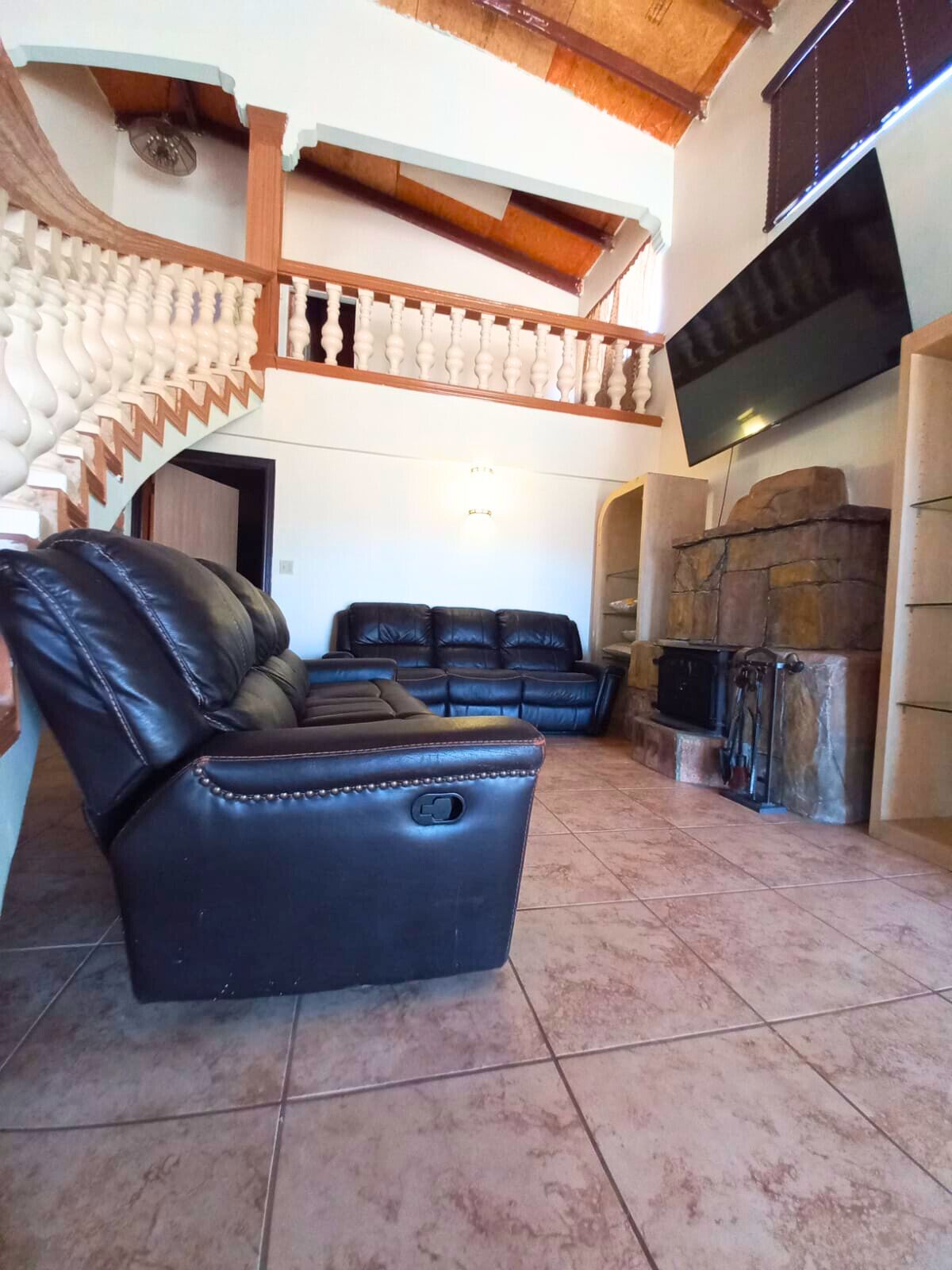 A spacious living area with a black leather sofa, a stone fireplace, and a staircase with a white balustrade.