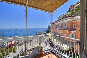 Ein Balkon mit Meer- und Bootsblick, ein überdachter Bereich mit Geländer sowie ein Gebäude mit Balkonen und Blumen.