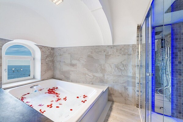 Ein modernes Badezimmer mit einer großzügigen Badewanne, einer Dusche mit Glastrennwand und Meerblick durch ein Fenster.