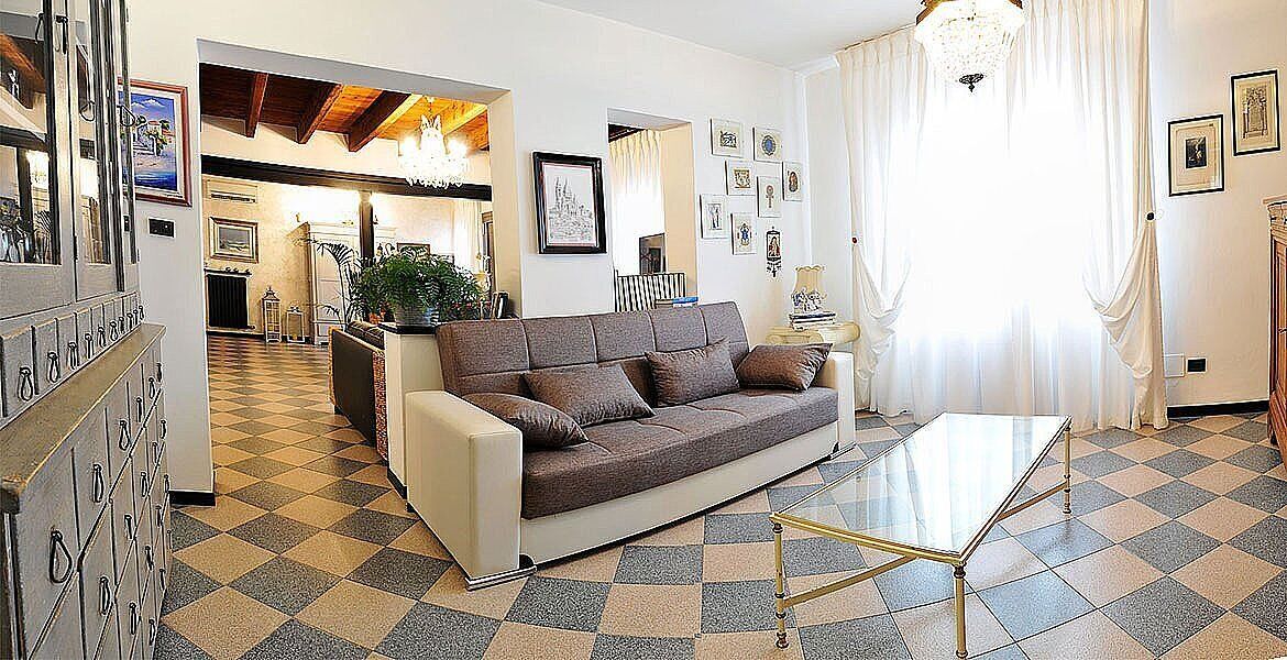 A living room with a sofa, a glass coffee table, and a checkered floor. There is a kitchen area in the background and a chandelier hanging from the ceiling.