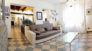 Ein Wohnzimmer mit einer Couch, einem Glastisch und einem Schachbrettfußboden. Im Hintergrund befindet sich eine Küchenzeile und eine Kristallleuchter hängt von der Decke.