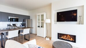 A modern living room with a fireplace, a kitchen area with grey cabinets and stainless steel appliances, and a flat-screen TV mounted above the fireplace.