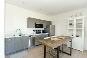 A modern kitchen with grey cabinets, stainless steel appliances, and a wooden dining table. - Executive Studio with Ocean & Mountain Views (Nanaimo)