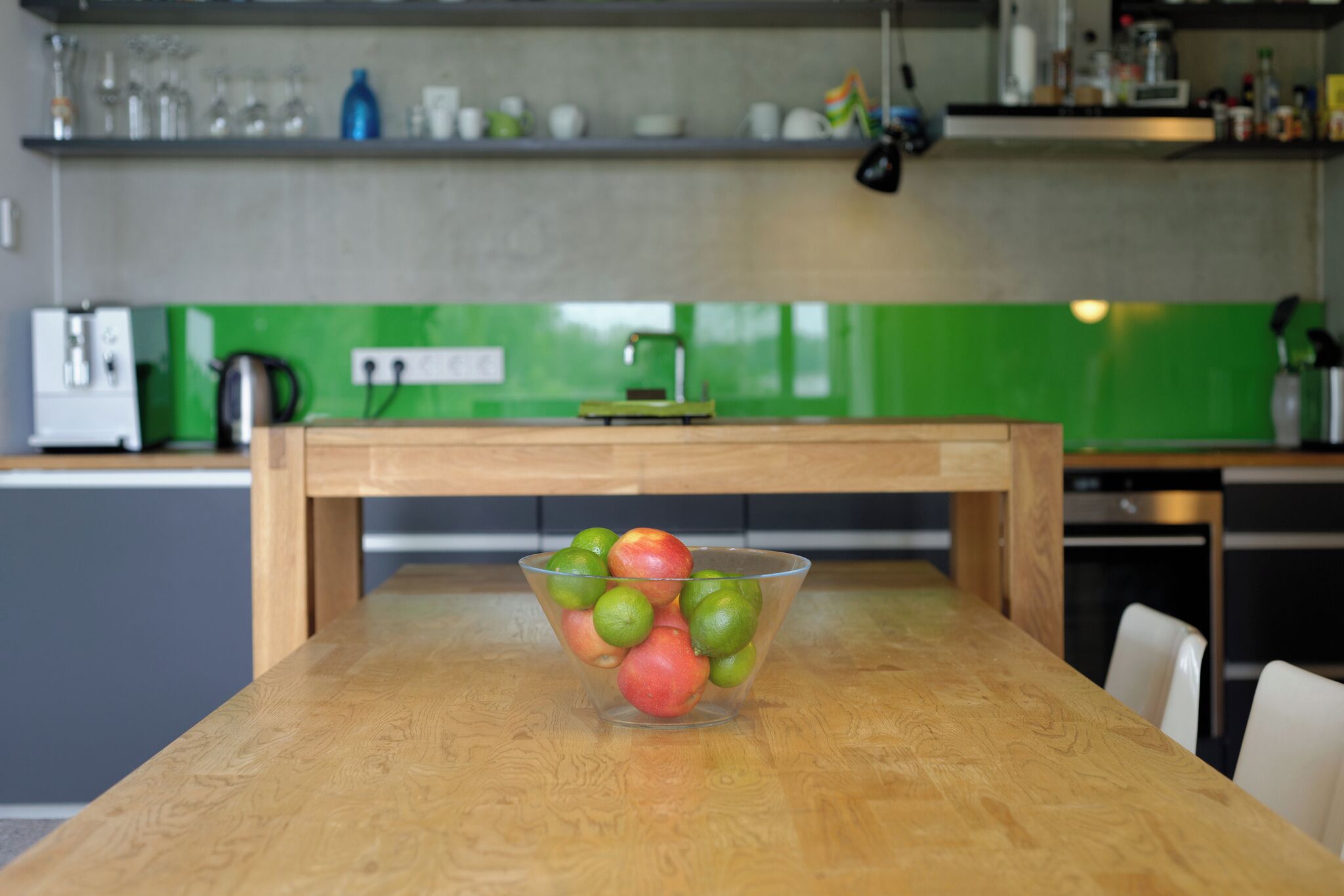 A kitchen with a wooden table, a bowl of apples and limes, and a green backsplash.