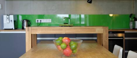 A kitchen with a wooden table, a bowl of apples and limes, and a green backsplash.