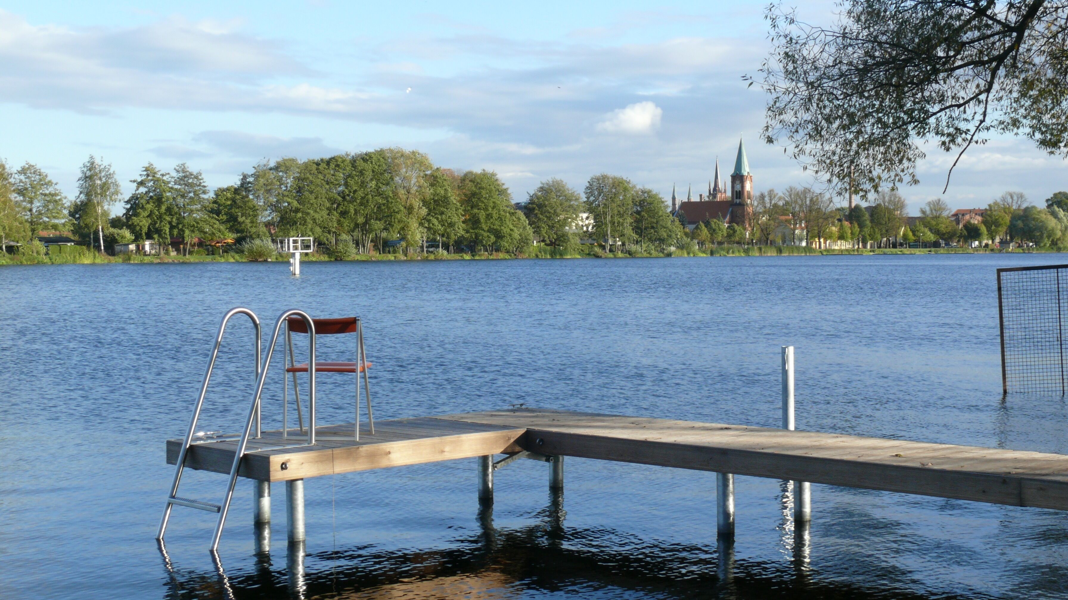 Ein Holzsteg führt in einen stillen See, mit einer Metallleiter und einem Stuhl. Im Hintergrund sieht man eine Kirche mit einem Kirchturm und eine Baumreihe.