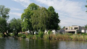 A lakeside view with a weeping willow tree, a dock, and modern houses in the background. - House on the Havelbogen directly on the water with canoe and jetty, terrace apartment***** (Werder (Havel))