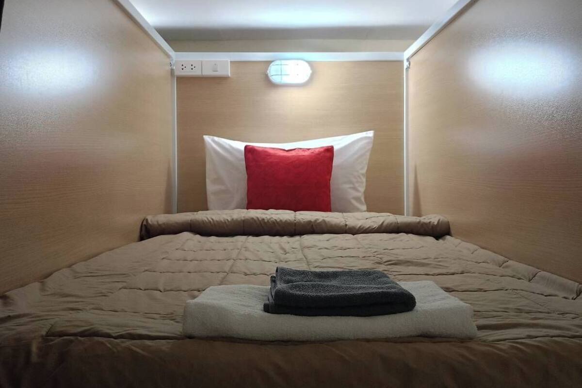 A small, cozy room with a bed, a red pillow, and a white pillow. There is a small window with a light fixture above the bed.