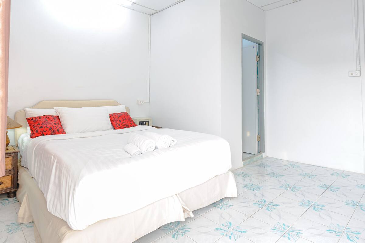 A neatly made bed with white and red pillows, a bedside table with a lamp, and a door leading to another room.