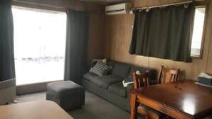 A living room with a wooden dining table, grey sofa, and a window with curtains. - Budget acc (Port Macdonnell)