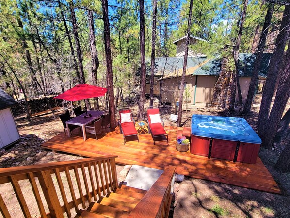 Una terraza de madera con mobiliario exterior, un jacuzzi y una pequeña construcción en zona boscosa.