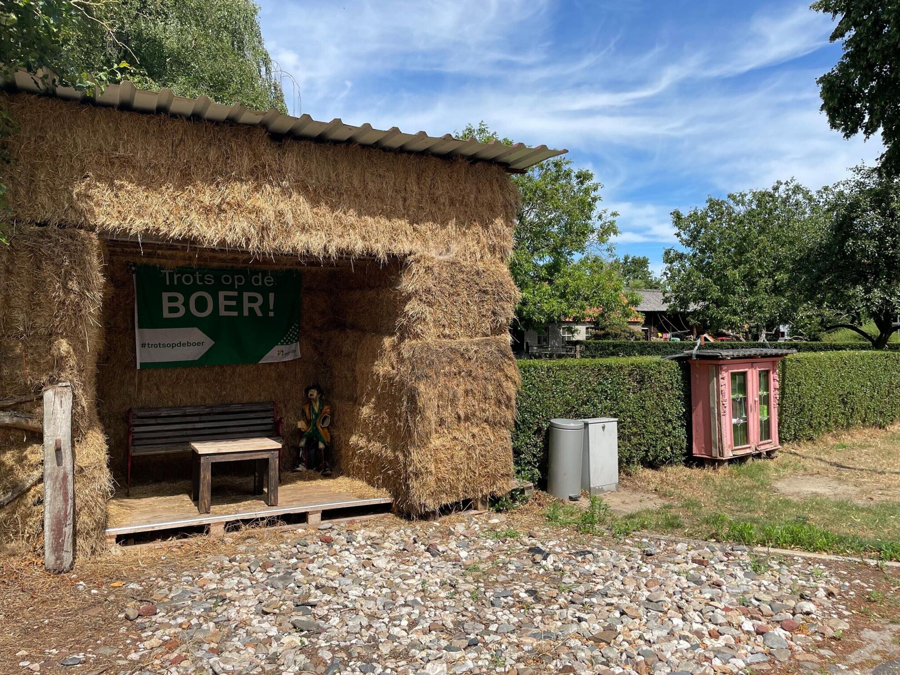 Un bâtiment rustique avec une enseigne verte sur laquelle on peut lire « Tots op de BOER ! » et un banc en bois à l’extérieur.