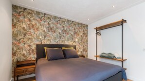 Une chambre à coucher comprenant un lit, une table de chevet, une étagère en bois et un mur recouvert d’un papier peint à motifs floraux.