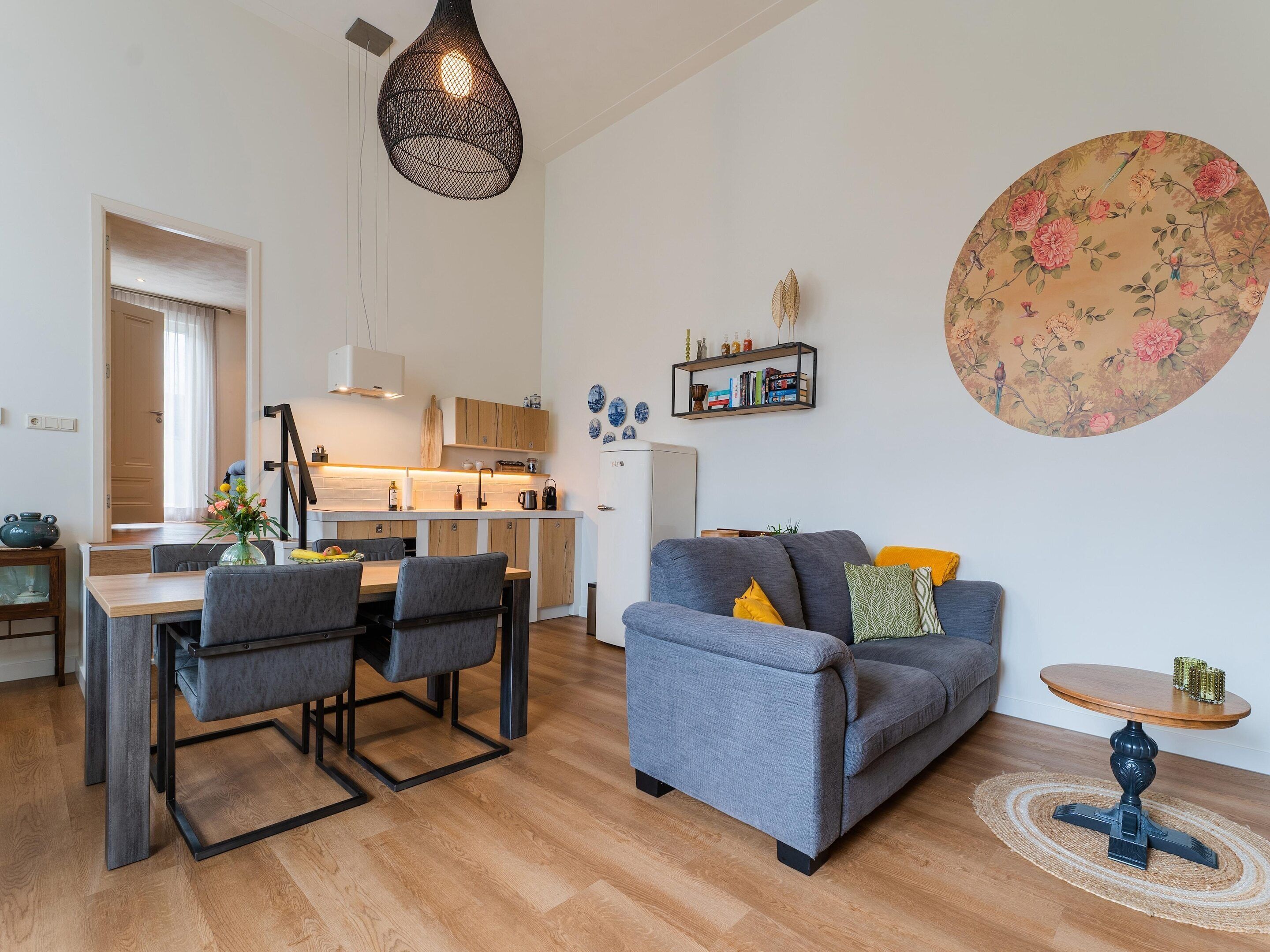 A modern living room with a grey sofa, a dining table with chairs, a kitchen area, and a large floral wall decoration.