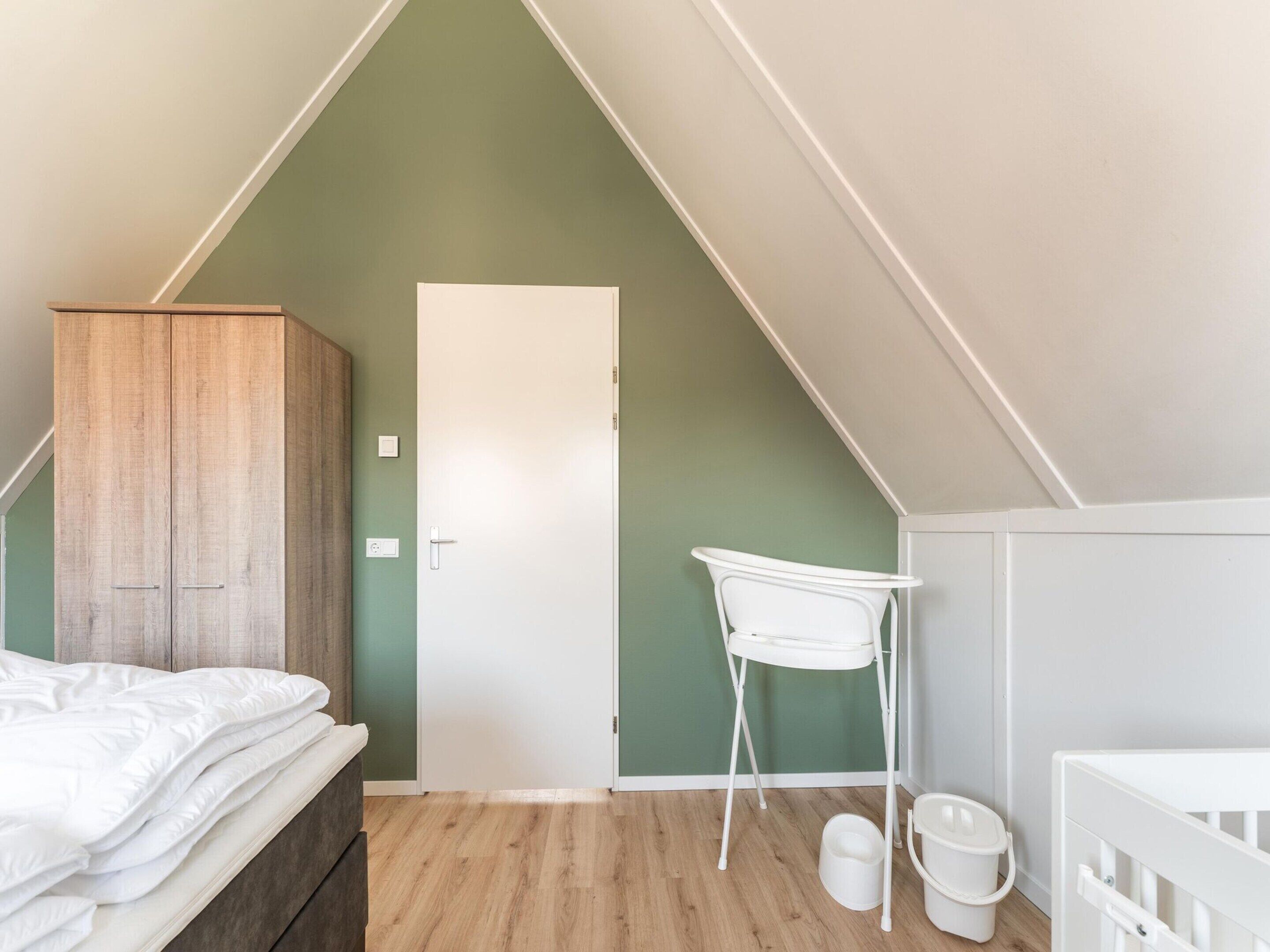 A bedroom with a wooden wardrobe, a white door, a white chair, and a crib.
