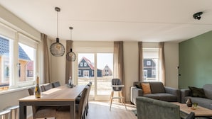 Ein geräumiger Wohnbereich mit Esstisch, Sofa und Sesseln. Große Fenster bieten Blick auf die Nachbarhäuser.