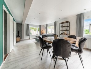 Un vaste salon doté d’une grande table à manger en bois, entourée de chaises modernes, d’une bibliothèque intégrée et offrant une vue sur l’extérieur grâce à de grandes fenêtres.