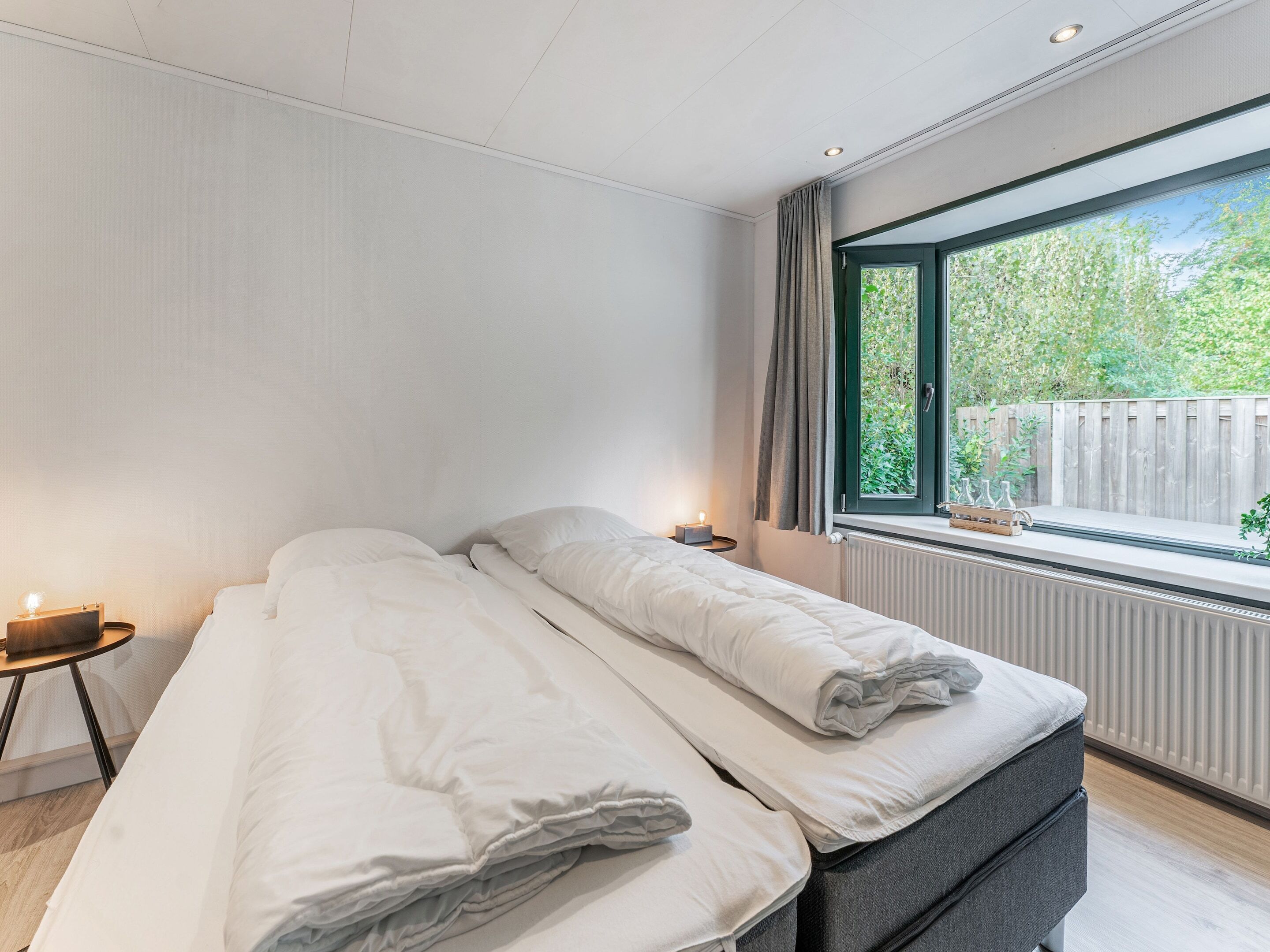 A modern bedroom with a bed, bedside table, and a window with a view of greenery.