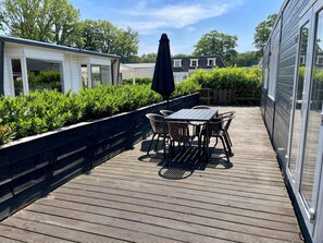 Une terrasse en bois avec une table et des chaises, entourée de verdure et dotée d’un espace abrité.