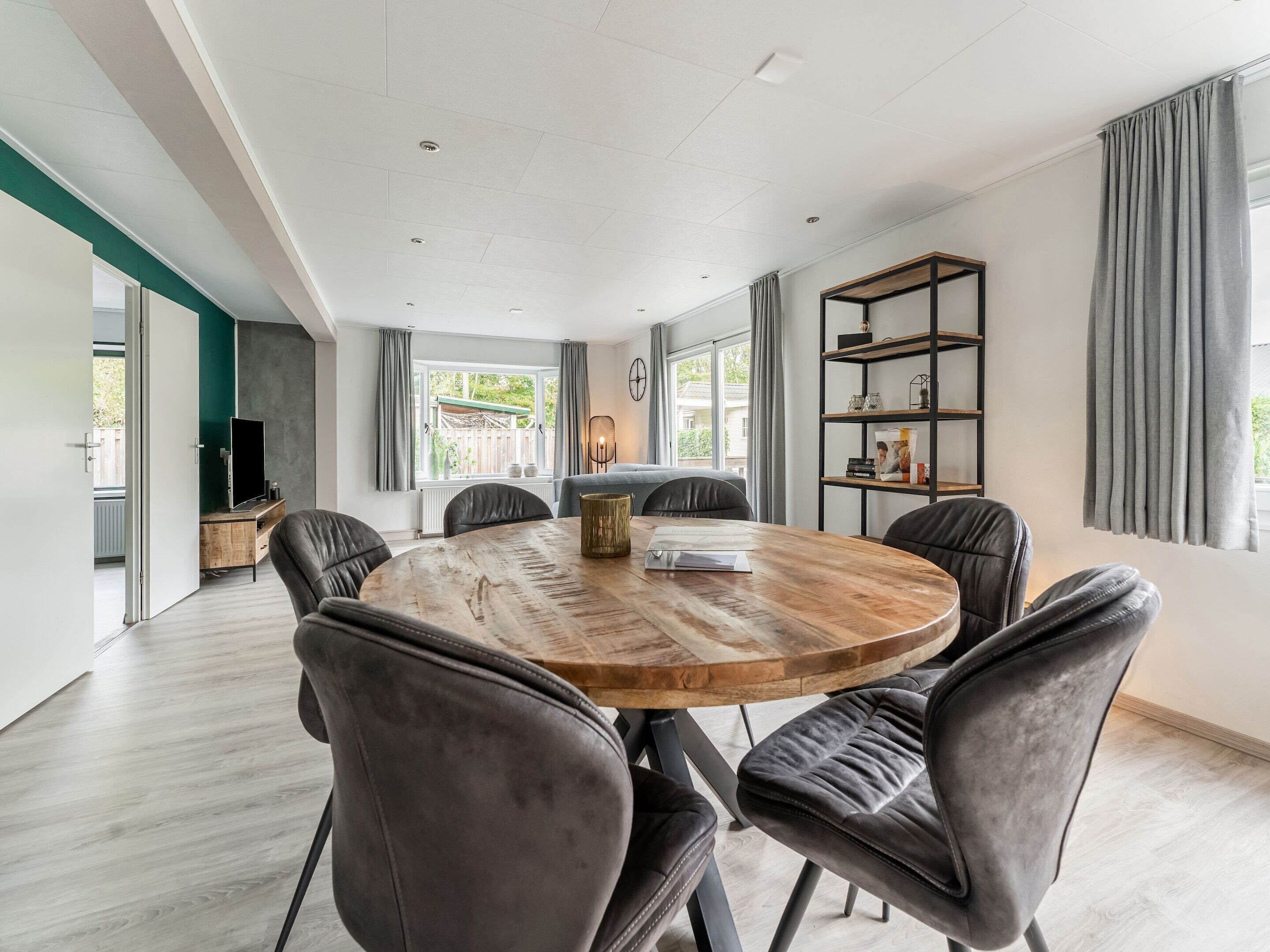 A modern dining area with a round wooden table, six upholstered chairs, a bookshelf, and a television on a stand.