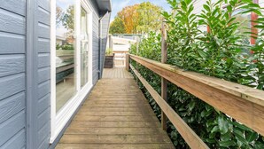 Una terraza de madera que lleva a una casa con puerta corrediza de vidrio, rodeada de follaje y un cielo despejado.