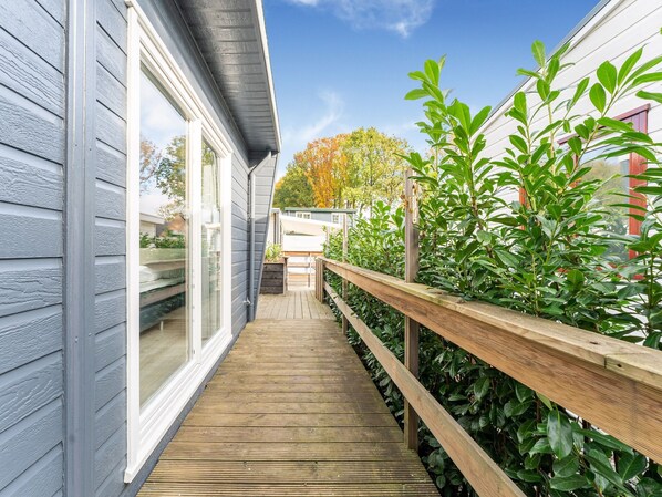 Une terrasse en bois menant à une maison dotée d’une porte-fenêtre coulissante, entourée de verdure et baignée d’un ciel dégagé.