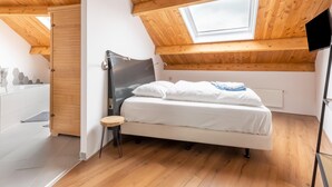 Een slaapkamer met dakraam, houten balken in het plafond, een bed met wit beddengoed en een klein krukje.