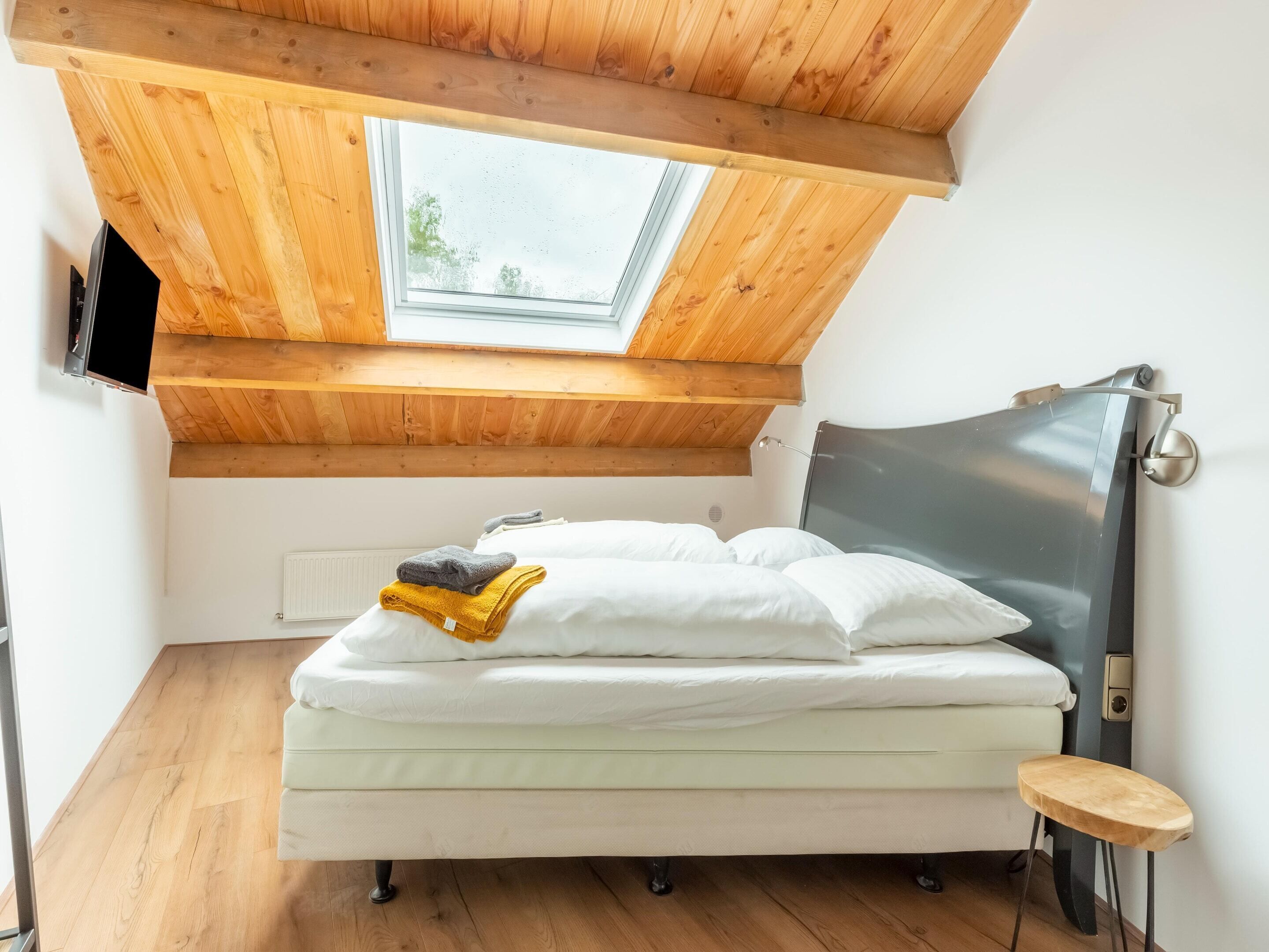Une chambre avec un plafond en pente, un puits de lumière, un lit avec une literie blanche, une table de chevet et une lampe fixée au mur.