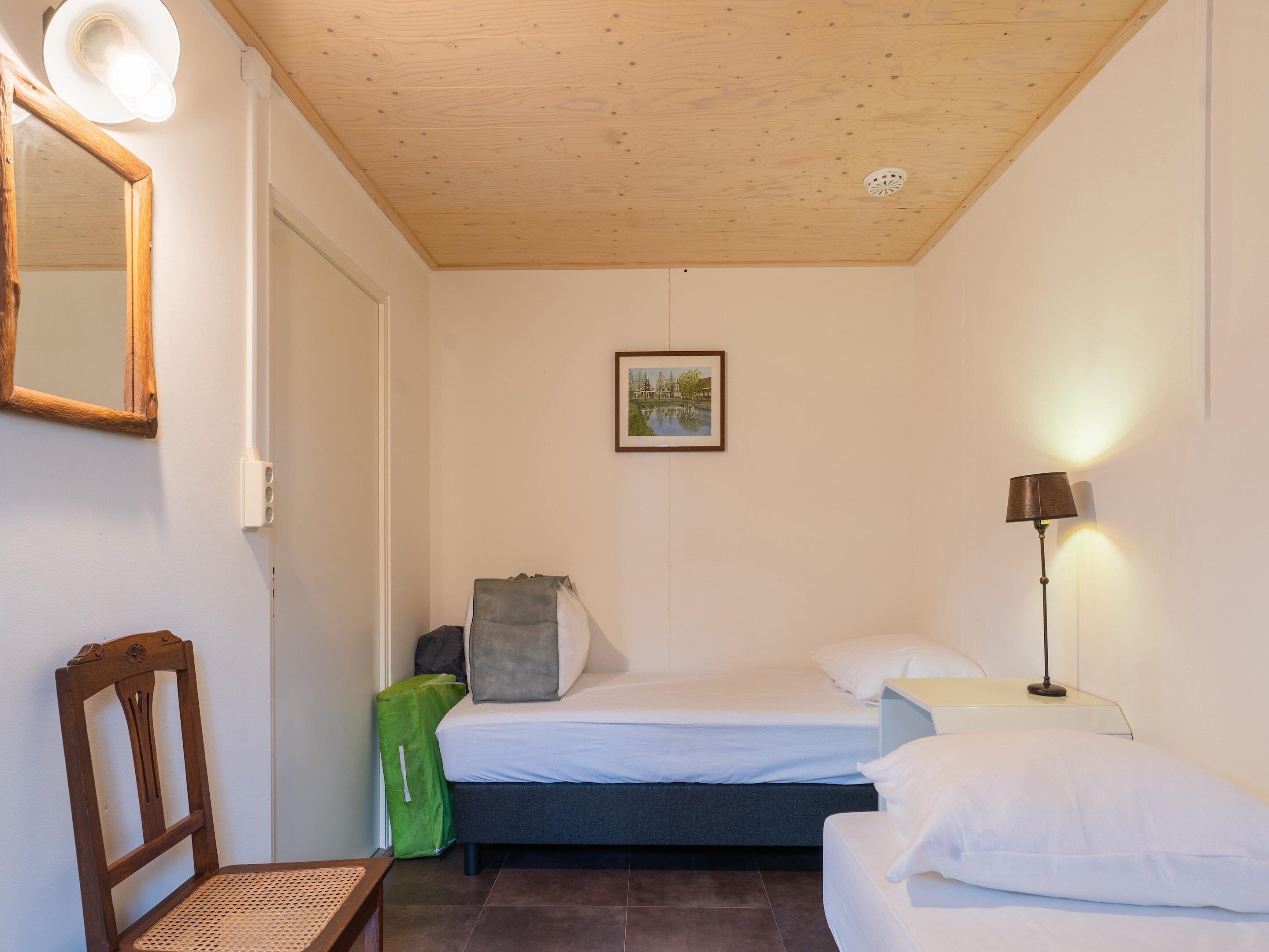 Et lille, rent værelse med en enkeltseng, en stol, en grøn taske og en lampe.